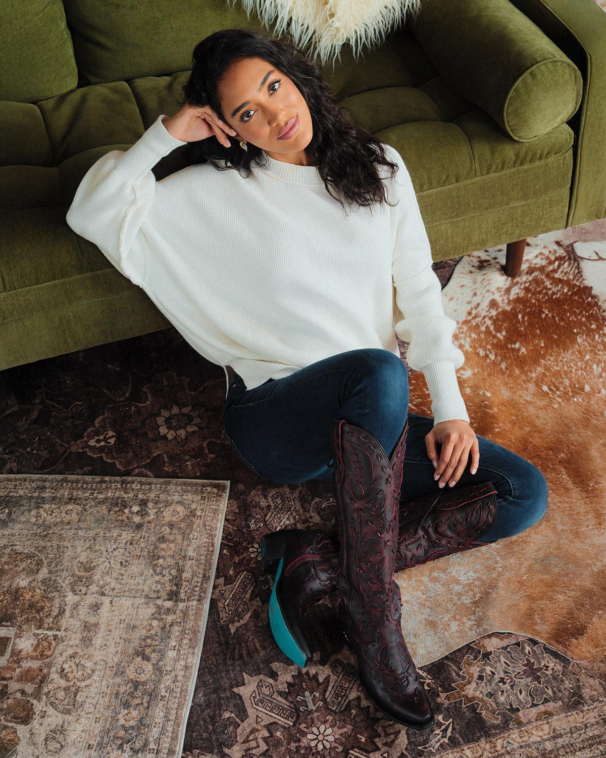 A woman wearing a white sweater, dark jeans, and tall maroon embroidered cowboy boots sits on the floor next to a green couch on a patterned rug