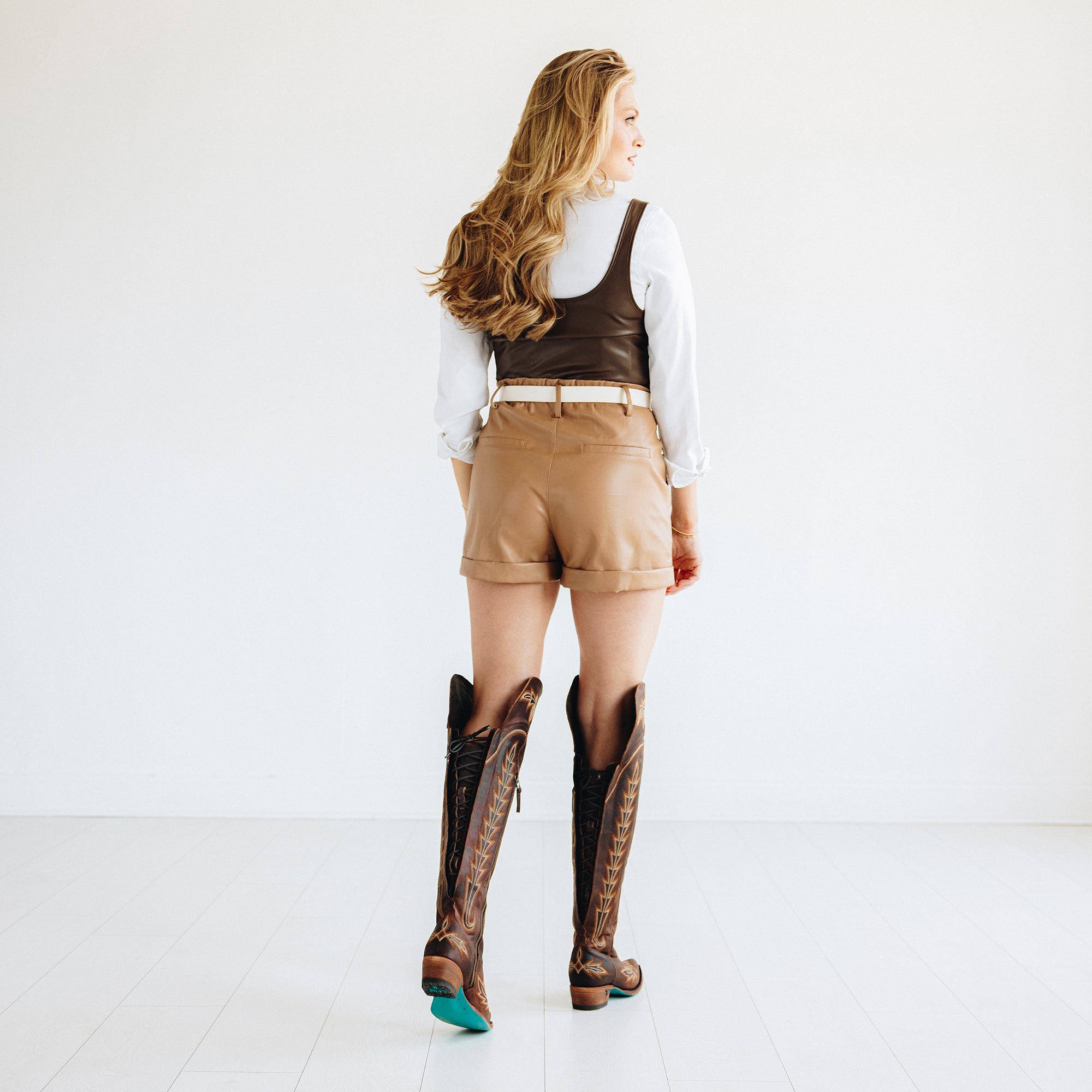 A woman standing with her back to the camera wears a white shirt, brown vest, tan shorts, and tall brown cowboy boots with turquoise soles