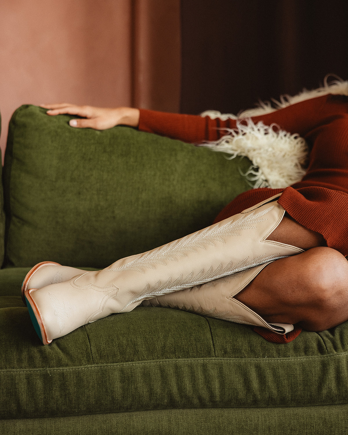 A woman reclining on a green couch wearing a rust-colored dress and tall cream cowboy boots with decorative stitching